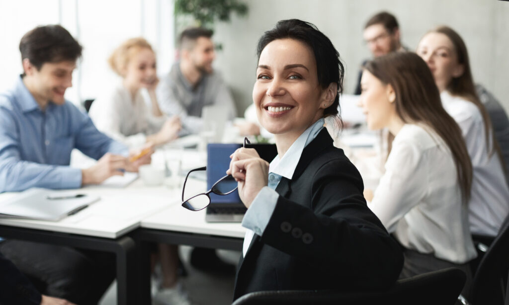 Top manager smiling to camera at meeting, colleagues discussing work on background, panorama